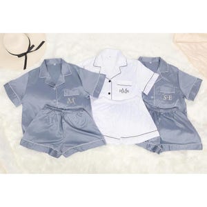 May include: Three sets of pyjamas in blue and white. The pyjamas have short sleeves and shorts. Each set has a different monogram embroidered on the pocket. The monograms are "S.E", "H.M.N", and "S.K".