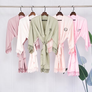 May include: Four satin robes with lace trim, hanging on hangers. The robes are in shades of pink, green, and white. Each robe has a different initial embroidered on the front.