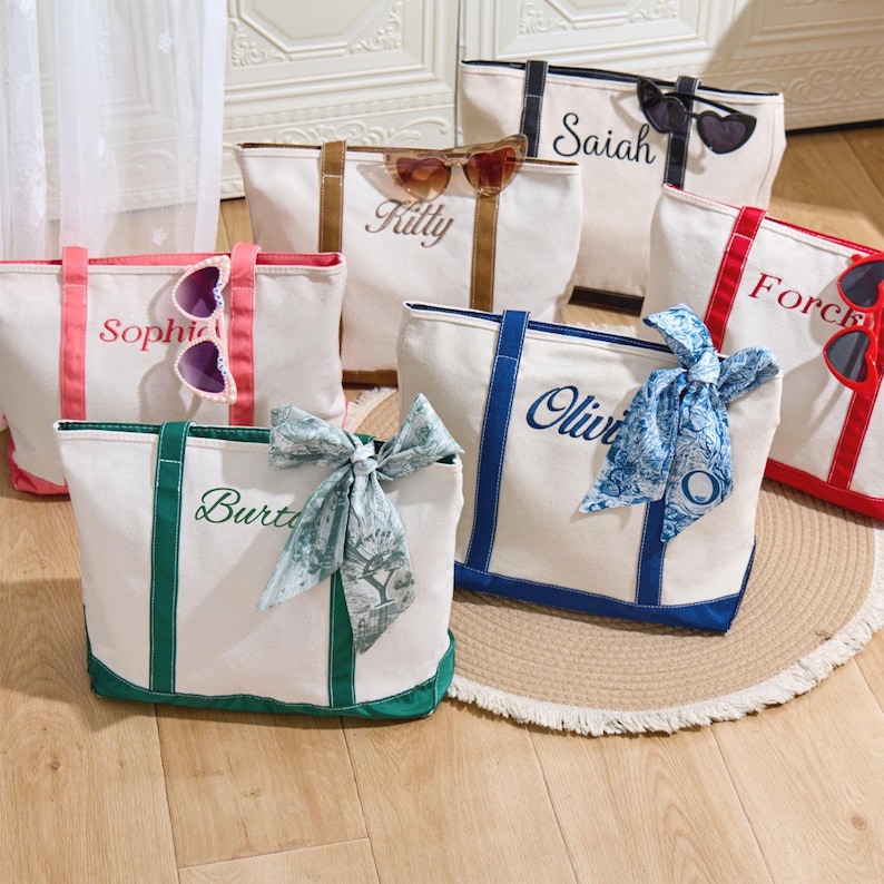 Puede incluir: Siete bolsas de lona blancas con nombres personalizados bordados en diferentes colores. Los nombres son: Sophia, Kitty, Saiah, Olivia, Burt, Force, y una s&eacute;ptima bolsa con una bufanda de patr&oacute;n azul y blanco.