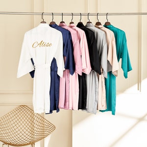 Könnte beinhalten: Eine Sammlung von zehn Satin-Bademänteln in verschiedenen Farben, darunter Weiß, Marineblau, Rosa, Schwarz, Grau, Beige und Türkis. Der weiße Bademantel hat den Namen "Alise" auf der Rückseite gestickt.