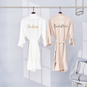 May include: Two white satin robes with ruffled hems, one with the word "Dulcie" embroidered in gold on the back, and the other with the words "Bridesmaid" embroidered in black on the back.