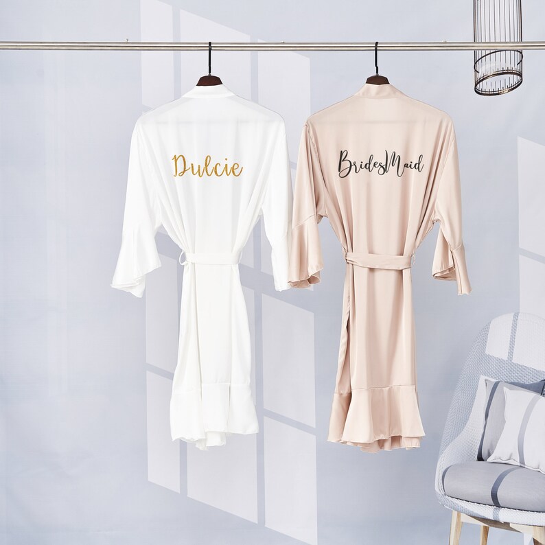 May include: Two white satin robes with ruffled hems, one with the word "Dulcie" embroidered in gold on the back, and the other with the words "Bridesmaid" embroidered in black on the back.