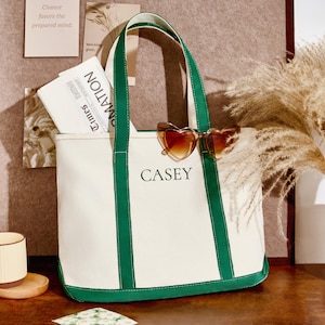 Könnte beinhalten: Eine weiße Canvas-Tragetasche mit grünen Verzierungen und Griffen, mit dem Namen "CASEY" in Schwarz. Die Tasche enthält eine Zeitung und herzförmige Sonnenbrillen. Eine Tasse und eine dekorative Vase sind ebenfalls zu sehen.