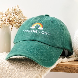 May include: A forest green baseball cap featuring an embroidered rainbow graphic above the white text "CUSTOM LOGO". The cap has a structured crown, a curved brim, and stitched eyelets on the top panels, showcasing a classic six-panel design.