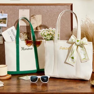 May include: Two canvas tote bags, one with green trim and the name "Moreno" embroidered in gold, the other with a floral ribbon and the name "Amanda". Both bags are off-white with long handles. Sunglasses and a coffee mug are on the table.