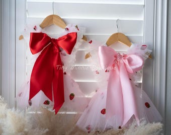May include: Two dresses with strawberry embroidery and large bows hang on wooden hangers. One dress has a red bow, the other a pink bow. The dresses are white with a tulle skirt and strawberry accents. The text "Tiny Minnie Boutique" is visible.