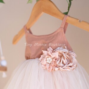 May include: A blush pink tulle dress with a blush pink bodice and a large blush pink flower with a pearl embellishment. The dress is hanging on a wooden hanger with the text "Tiny Minnie Boutique" on the bodice.
