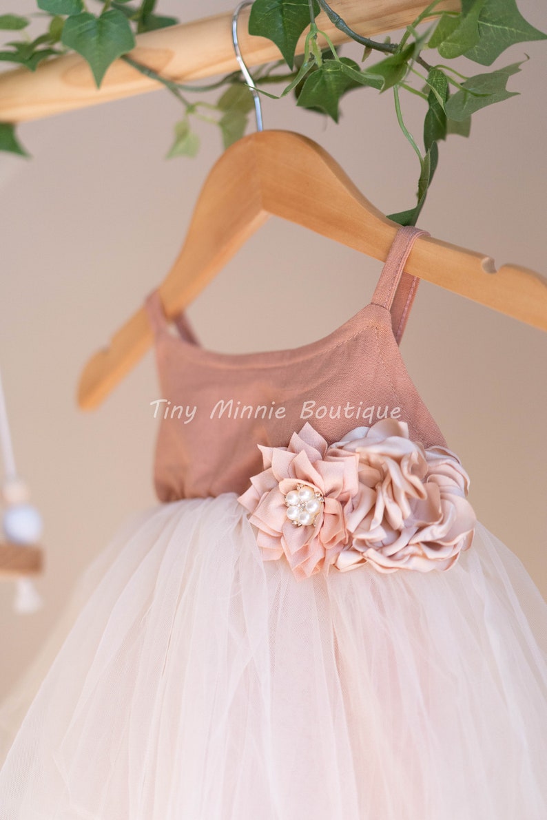 May include: A blush pink tulle dress with a blush pink bodice and a large blush pink flower with a pearl embellishment. The dress is hanging on a wooden hanger with the text "Tiny Minnie Boutique" on the bodice.