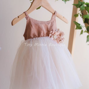 May include: A blush pink tulle dress with a brown bodice and a flower embellishment. The dress is hanging on a wooden hanger. "Tiny Minnie Boutique" is written on the dress.