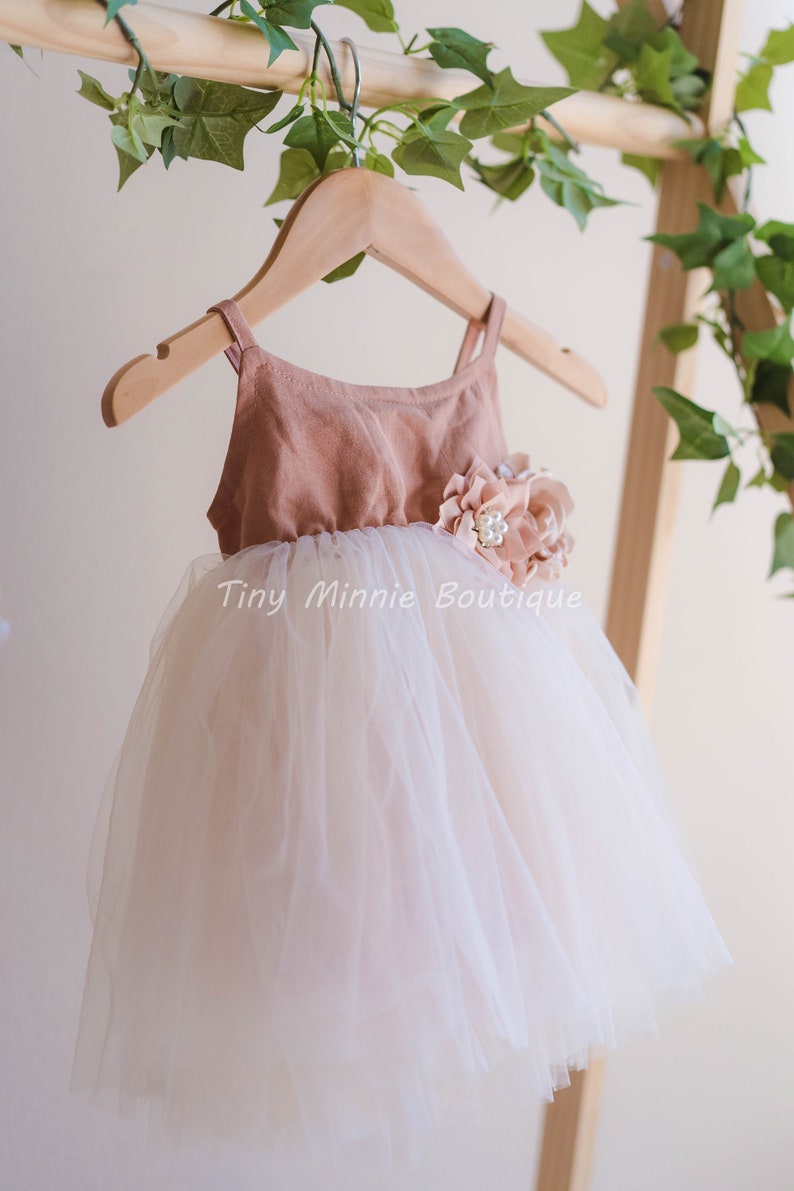 May include: A blush pink tulle dress with a brown bodice and a flower embellishment. The dress is hanging on a wooden hanger. "Tiny Minnie Boutique" is written on the dress.