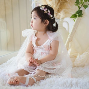 May include: A young girl wearing a white dress with pink butterfly accents and a flower crown. She is sitting on a white fluffy rug and looking to the side.