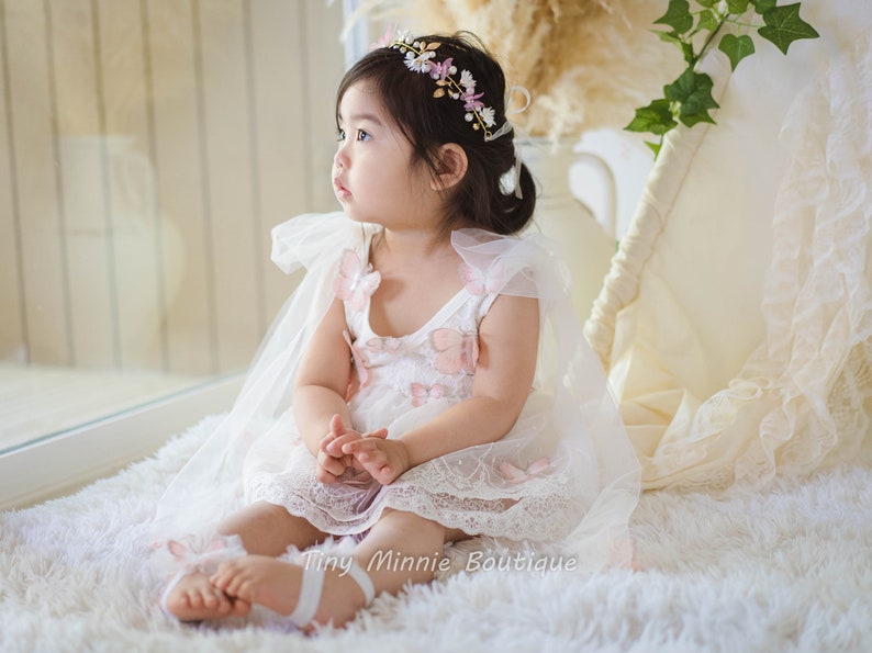 May include: A young girl wearing a white dress with pink butterfly accents and a flower crown. She is sitting on a white fluffy rug and looking to the side.