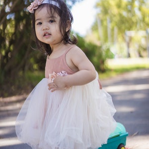 May include: A young girl wearing a white tulle dress with a pink bodice and a flower headband. She is standing on a paved path with a toy wagon behind her.