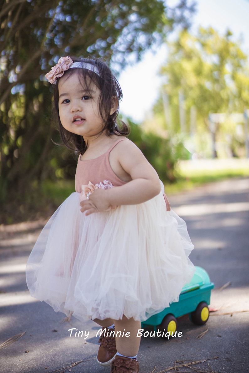 May include: A young girl wearing a white tulle dress with a pink bodice and a flower headband. She is standing on a paved path with a toy wagon behind her.