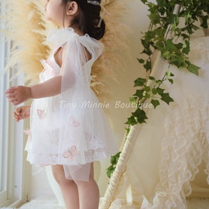 May include: A young girl wearing a white lace dress with pink butterfly appliques and a flower crown. She is standing on a white fluffy rug and looking up.