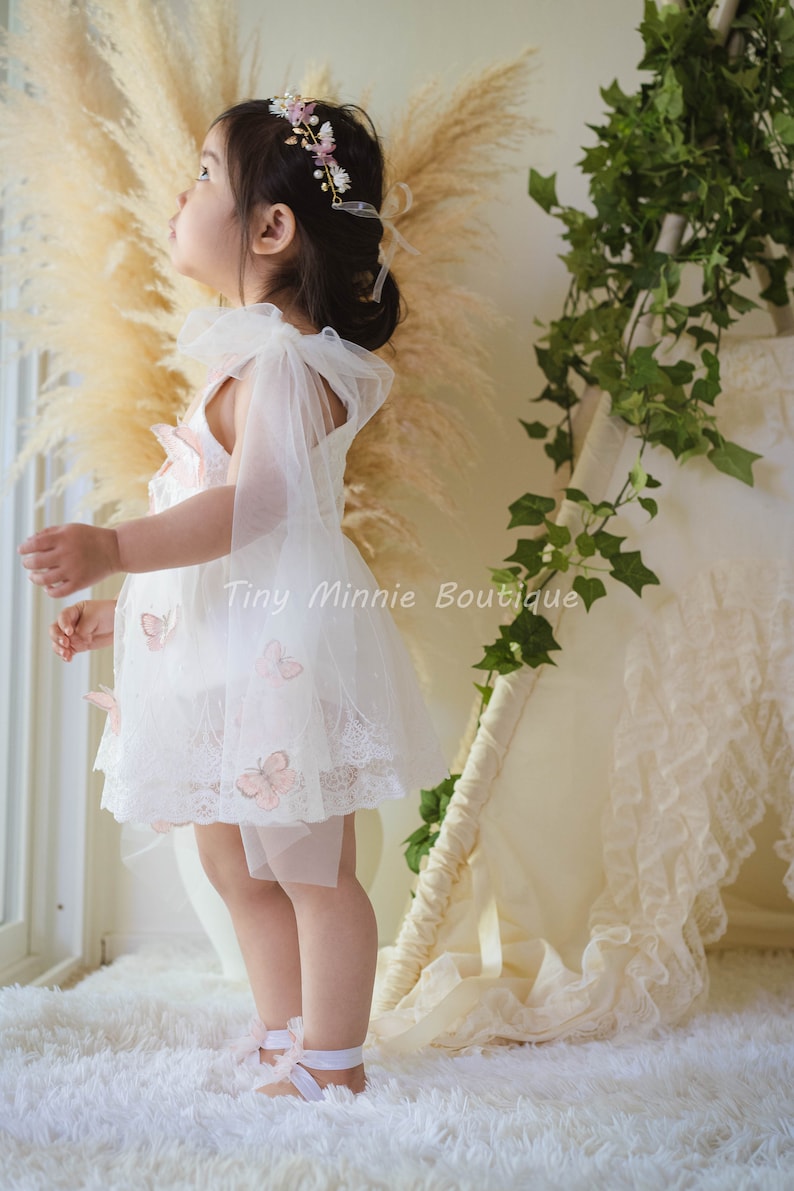 May include: A young girl wearing a white lace dress with pink butterfly appliques and a flower crown. She is standing on a white fluffy rug and looking up.