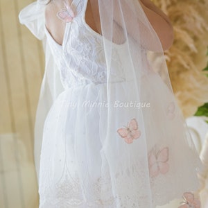 May include: White tulle dress with a lace bodice and butterfly embroidery. The dress has a large bow on the shoulder and a scalloped lace hem.