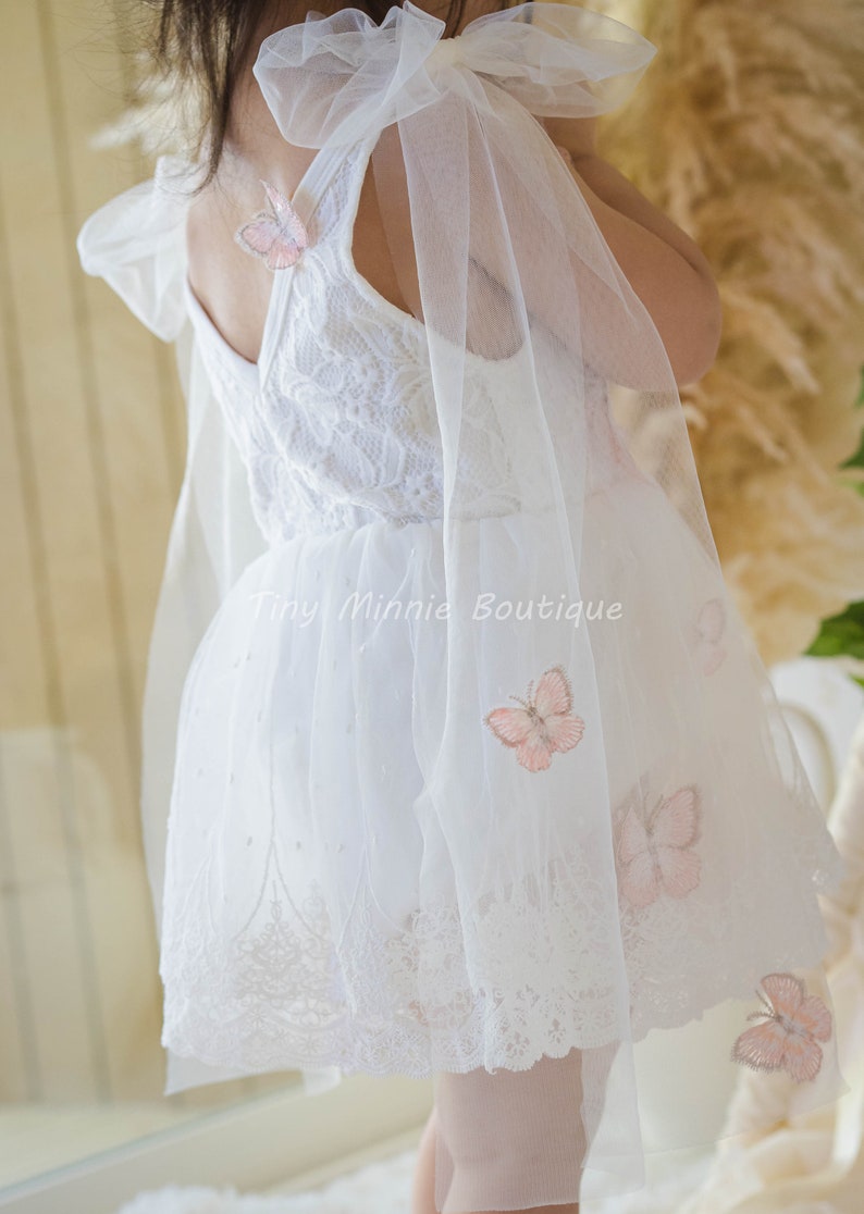 May include: White tulle dress with a lace bodice and butterfly embroidery. The dress has a large bow on the shoulder and a scalloped lace hem.