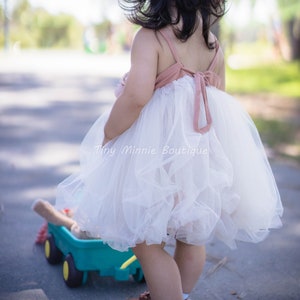 May include: A young girl wearing a white tulle dress with a pink bodice and a bow in the back. The dress is layered and has a ruffled hem. The girl is standing in a park and is holding a toy wagon.