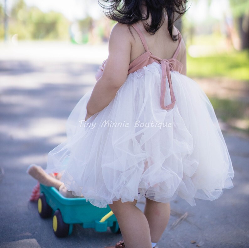 May include: A young girl wearing a white tulle dress with a pink bodice and a bow in the back. The dress is layered and has a ruffled hem. The girl is standing in a park and is holding a toy wagon.