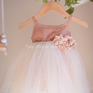 May include: A blush pink tulle dress with a matching bodice and a large flower embellishment. The dress is on a wooden hanger.