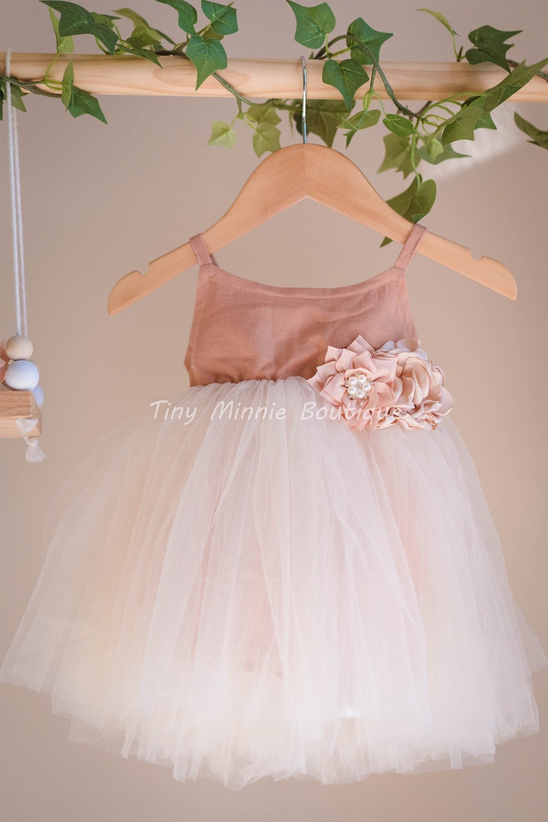 May include: A blush pink tulle dress with a matching bodice and a large flower embellishment. The dress is on a wooden hanger.
