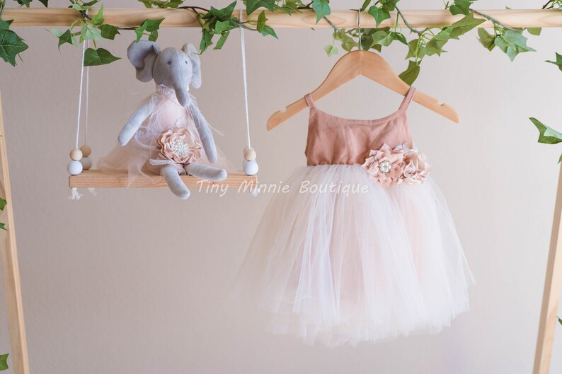 May include: A blush pink tulle dress with a brown bodice and a flower embellishment. The dress is hanging on a wooden hanger. The text "Tiny Minnie Boutique" is visible in the image.