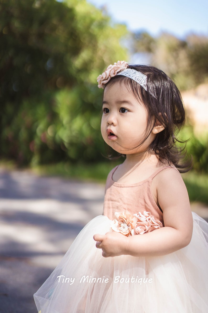 May include: A young girl wearing a white tulle dress with a pink bodice and a flower headband. The dress has a pink flower embellishment on the bodice. The girl is looking to the right of the frame. The text "Tiny Minnie Boutique" is visible at the bottom of the image.