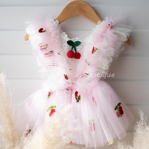 May include: A light pink tulle dress featuring ruffled shoulder straps and embroidered red cherry motifs with green stems. The bodice includes a white lace trim detail and the text TinyBunnieBoutique is visible across the center of the garment.