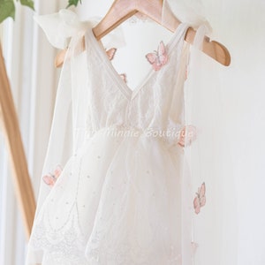 May include: A white tulle dress with a lace trim and pink butterfly appliques. The dress has a V-neckline and is hanging on a wooden hanger. The text "Tiny Minnie Boutique" is visible on the dress.