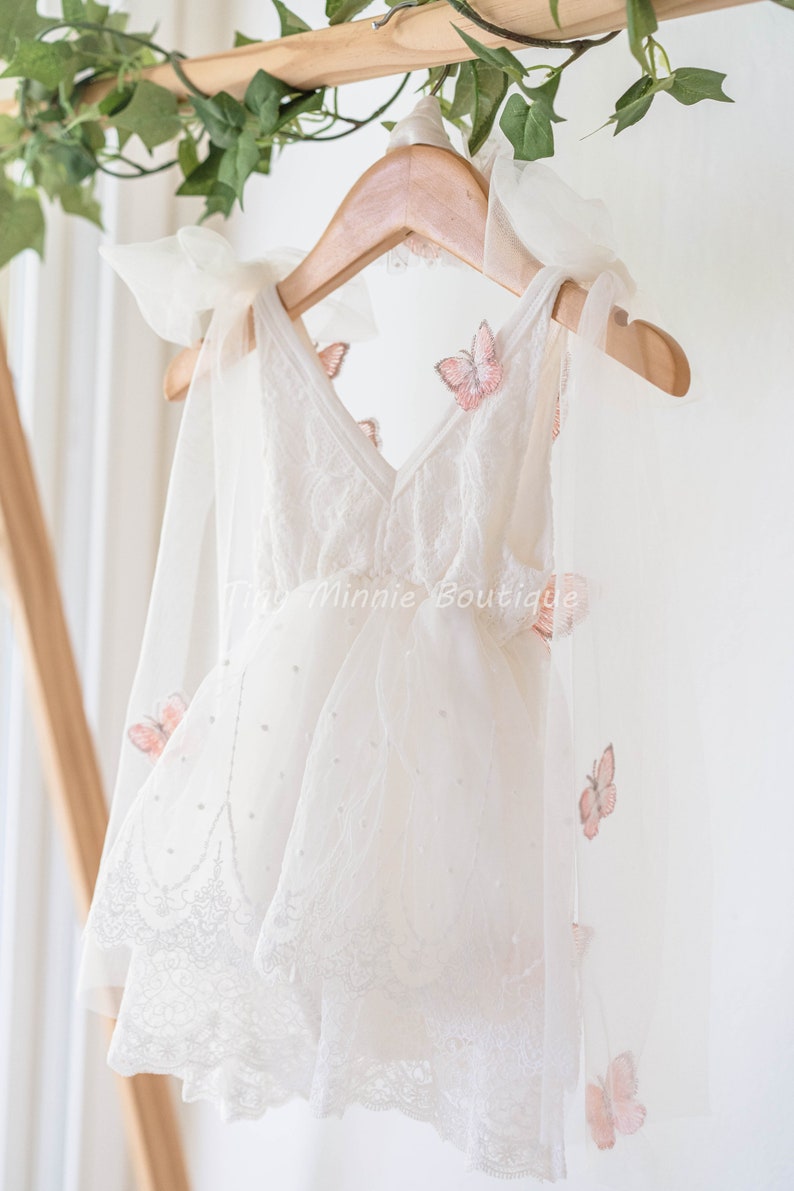 May include: A white tulle dress with a lace trim and pink butterfly appliques. The dress has a V-neckline and is hanging on a wooden hanger. The text "Tiny Minnie Boutique" is visible on the dress.