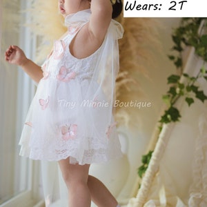 May include: A young girl wearing a white lace dress with pink butterfly embellishments and a flower crown. The dress has a tulle overlay and the girl is wearing white shoes. The text "BABY MODEL" is written above the girl's head, along with her weight, height, and size. The text "Tiny Minnie Boutique" is written below the girl.