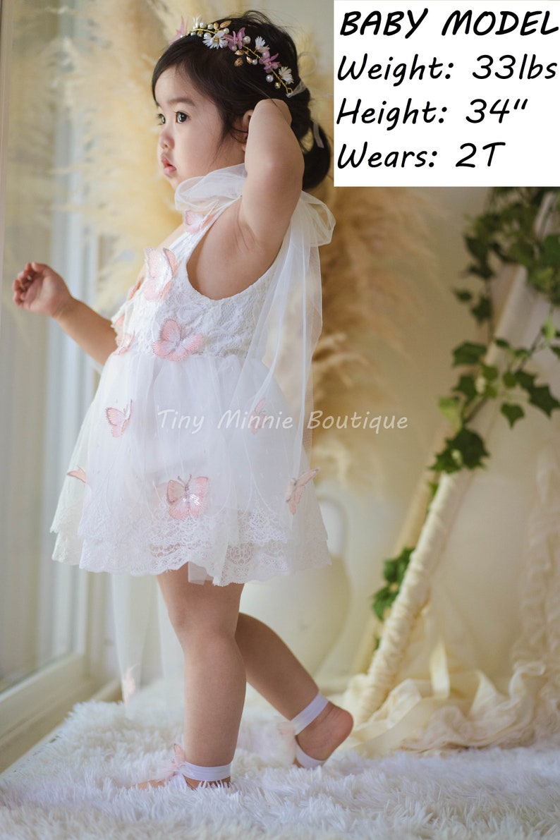 May include: A young girl wearing a white lace dress with pink butterfly embellishments and a flower crown. The dress has a tulle overlay and the girl is wearing white shoes. The text "BABY MODEL" is written above the girl's head, along with her weight, height, and size. The text "Tiny Minnie Boutique" is written below the girl.