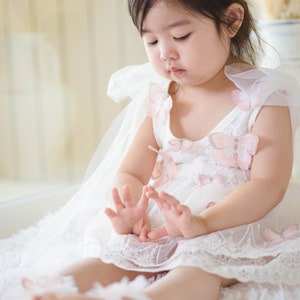 May include: A young girl wearing a white dress with pink butterfly embellishments and a gold floral crown. She is sitting on a white fluffy surface. The dress has a tulle overlay and the girl is wearing white shoes.