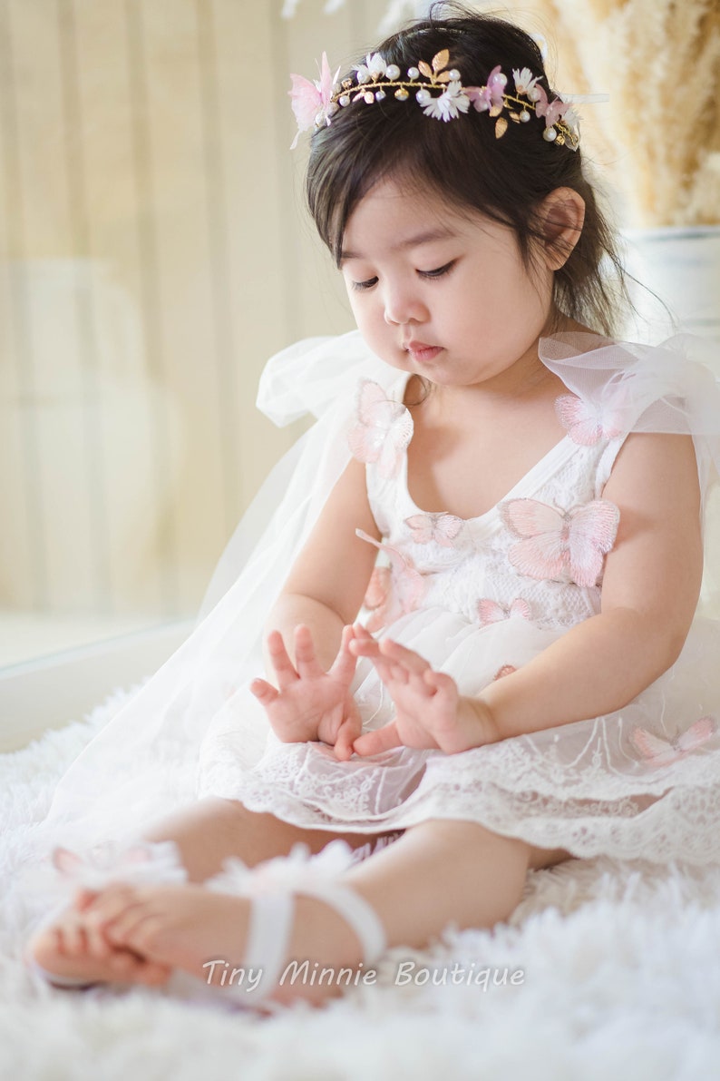 May include: A young girl wearing a white dress with pink butterfly embellishments and a gold floral crown. She is sitting on a white fluffy surface. The dress has a tulle overlay and the girl is wearing white shoes.