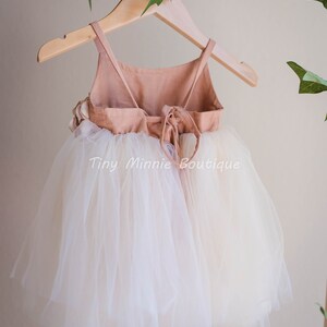 May include: A white tulle dress with a peach-colored bodice and a tie back. The dress is hanging on a wooden hanger with green leaves in the background. The text "Tiny Minnie Boutique" is visible on the dress.