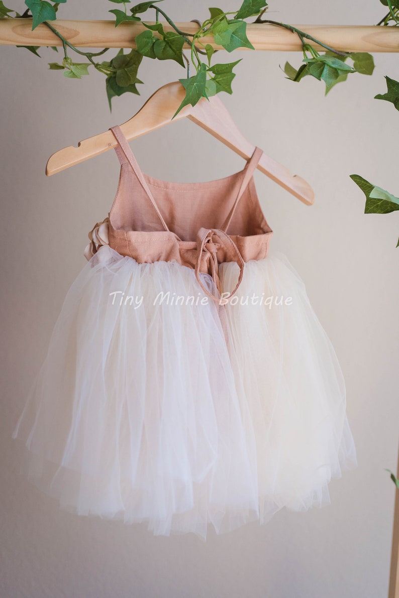 May include: A white tulle dress with a peach-colored bodice and a tie back. The dress is hanging on a wooden hanger with green leaves in the background. The text "Tiny Minnie Boutique" is visible on the dress.