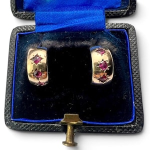 May include: A pair of gold hoop earrings featuring three round, deep pink rubies set into each earring. The rubies are surrounded by star-shaped engravings on the polished gold surface. The earrings are displayed inside a black jewellery box.