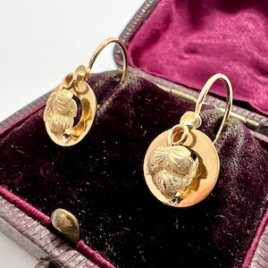 May include: A pair of gold earrings with a detailed floral design. The earrings are hanging from a velvet box.