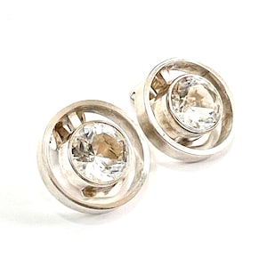 May include: A pair of silver earrings with a clear crystal stone set in a circular frame.