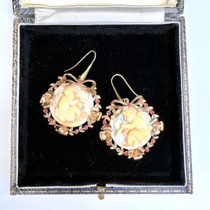 May include: A pair of gold tone earrings with a cameo of a cherub in a wreath of gold leaves and red stones.
