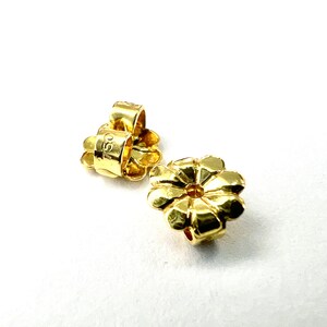 以下が含まれることがあります： 金色のイヤリングのバック。バックは花の形をしていて、中央に小さな穴が開いています。バックには「925」という数字が刻印されています。