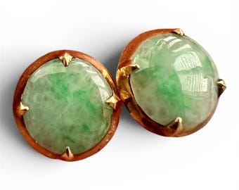 Könnte beinhalten: Ein Paar runde Ohrstecker mit polierten, durchscheinenden grünen Jade-Cabochons in warmfarbenen Goldmetallfassungen. Jeder kreisförmige Stein weist natürliche, gesprenkelte grüne Muster auf und wird von vier kleinen, spitzen Goldkrallen gehalten.