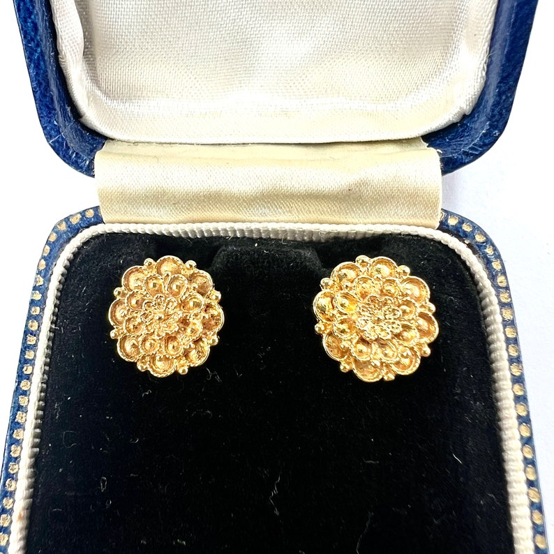 以下が含まれることがあります： 花柄のデザインの金色のイヤリングのペア。イヤリングは青いベルベットのジュエリーボックスに入っています。