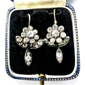 May include: A pair of antique earrings with a floral design, set in silver. Each earring features a cluster of clear and champagne-colored gemstones, with a teardrop-shaped dangle. The earrings are displayed in a blue and black jewelry box.