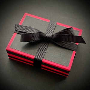 K&ouml;nnte beinhalten: Eine schwarze Geschenkbox mit rotem Rand und einem schwarzen Satinband, das zu einer Schleife gebunden ist.