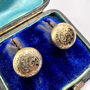 Puede incluir: Un par de gemelos redondos de color dorado con intrincados diseños florales grabados. Los gemelos se exhiben en un joyero vintage de terciopelo azul abierto, con exterior negro y forro interior turquesa.