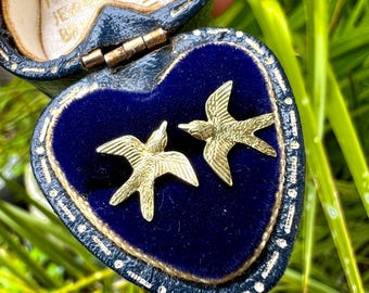 Könnte beinhalten: Ein Paar goldfarbene, vogelformige Ohrringe, präsentiert in einer herzförmigen, marineblauen Samt-Schmuckschatulle. Die Schatulle hat goldfarbene Verzierungen und einen goldenen Verschluss. Die Ohrringe sind mit Flügeln und Schnäbeln versehen.
