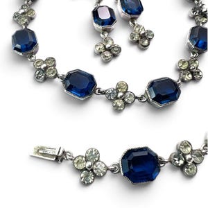Op de afbeelding: Een sieradenset bestaande uit een ketting en bijpassende oorbellen. De stukken zijn voorzien van grote, achthoekige, diepblauwe gefacetteerde stenen die afwisselen met kleine, vierbladige bloemen van heldere, sprankelende witte kristallen.