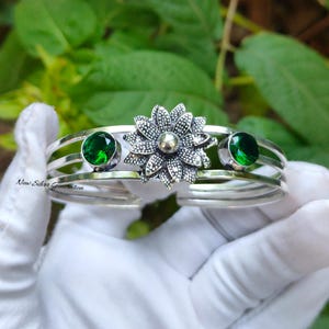 May include: Silver cuff bracelet featuring a central flower design and two round, green gemstones. The bracelet has three parallel bands and is displayed against a green foliage background. The text "New Silver Generation" is visible.